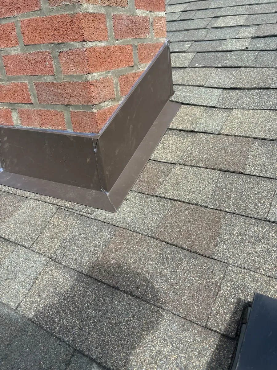 A new metal saddle and flashing fitted behind a brick chimney on a shingle roof
