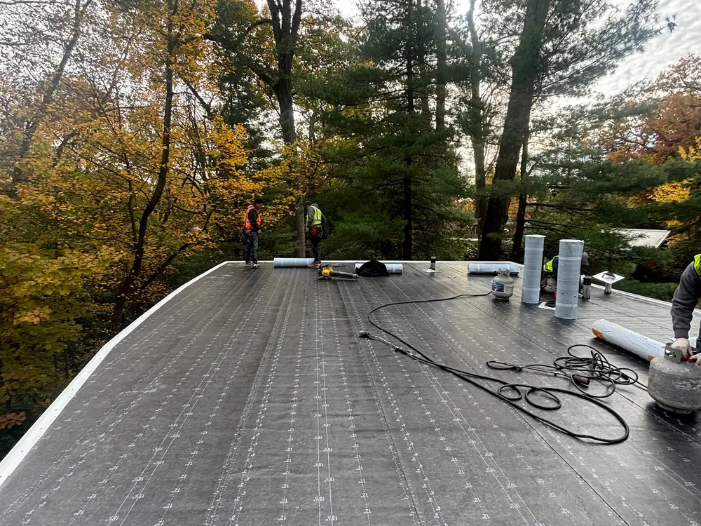 A flat roof mid installation with rolls and tools laid out, autumn trees behind