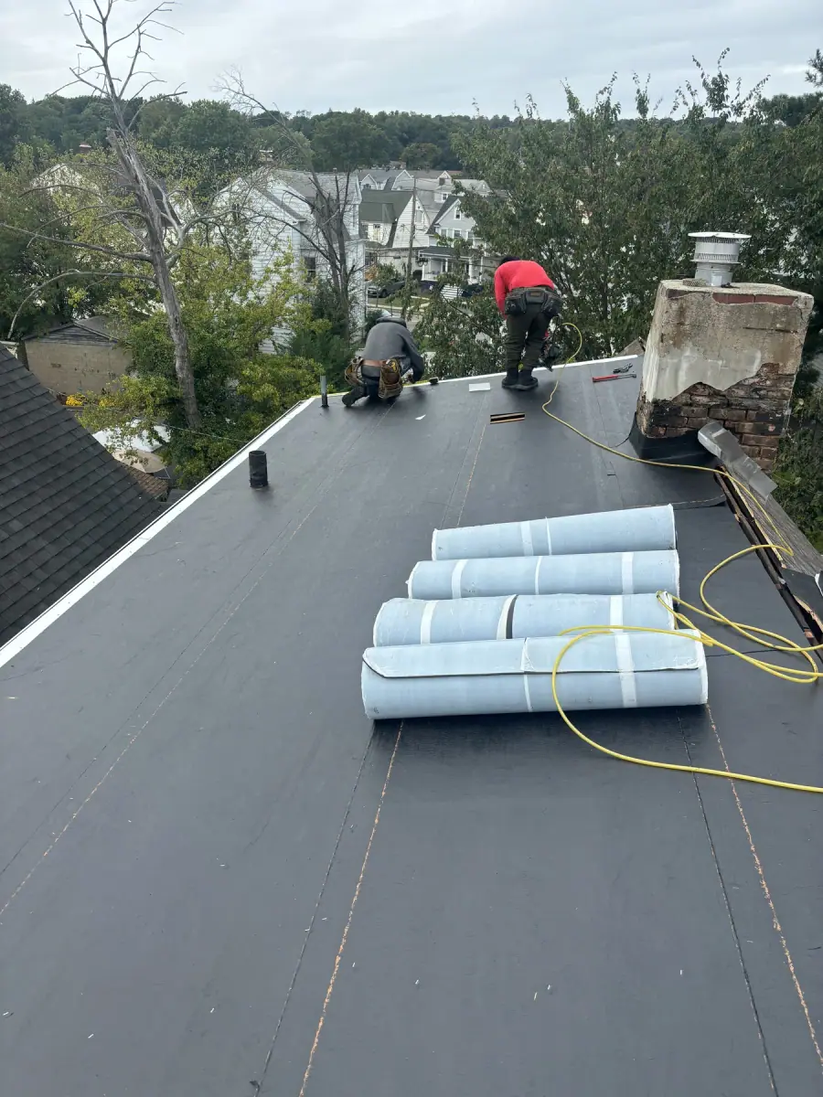 A long flat roof finished in black membrane with rolls staged and two crew at the far end