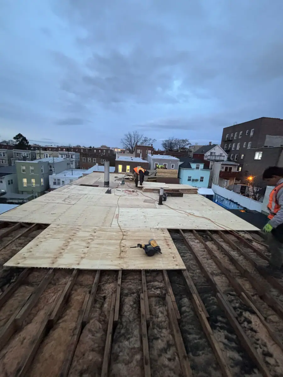 New plywood being laid over old plank decking on a flat roof at dusk, two crew working