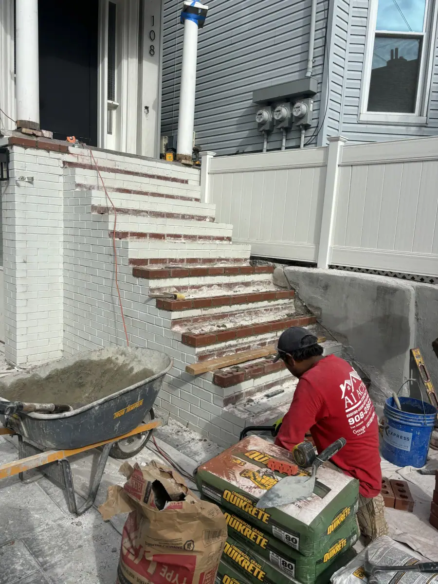 A crew member in a red A1 Action shirt laying new brick on front steps, a barrow of mortar beside him
