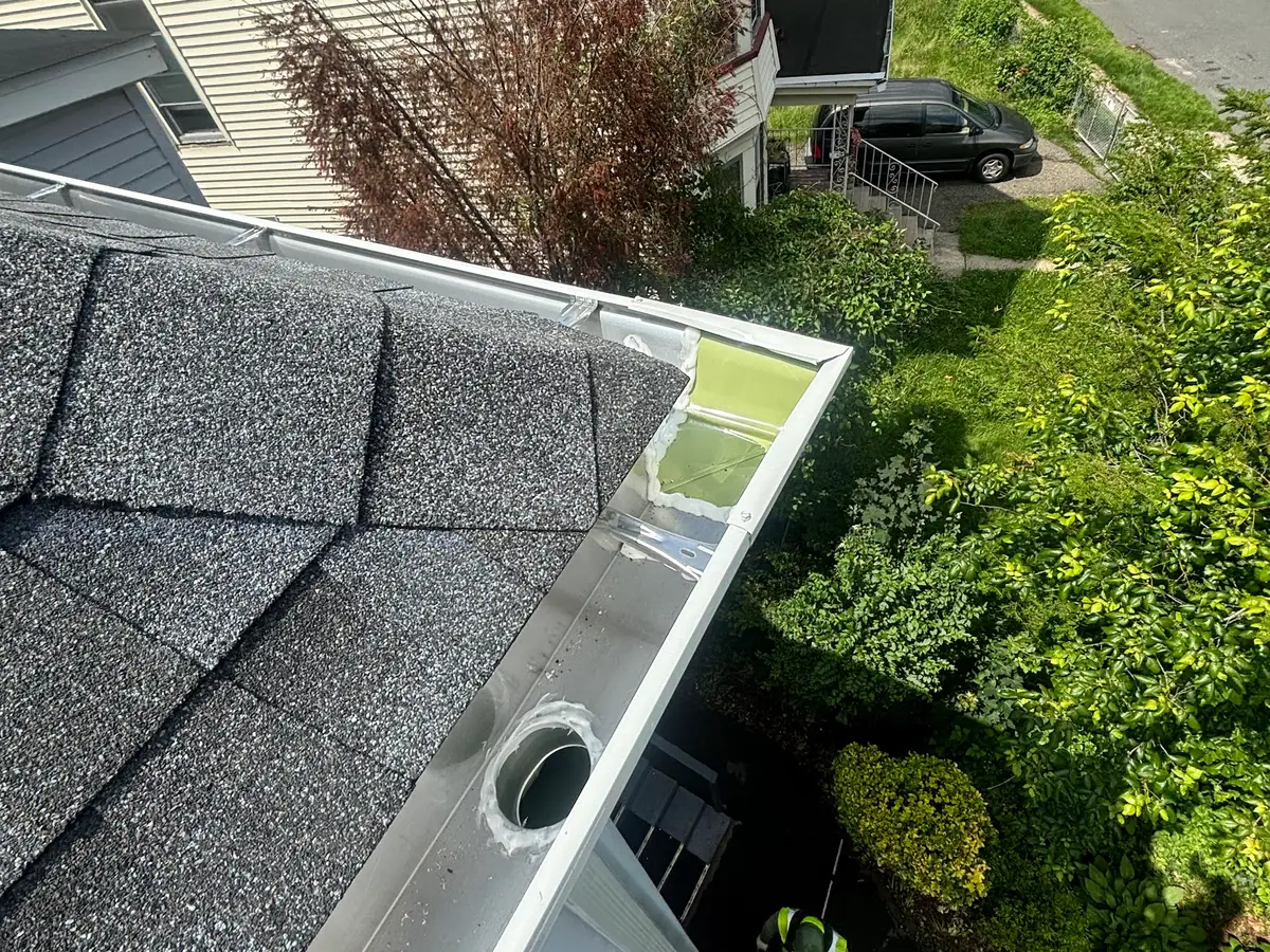 A new white gutter and outlet being fitted along the edge of a shingle roof