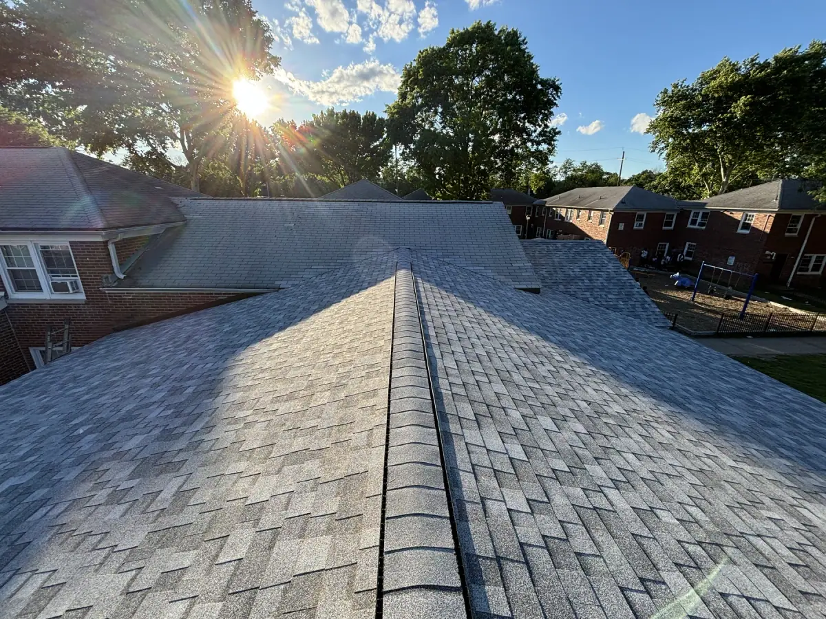 The ridge of a finished shingle roof with the sun flaring over the trees behind