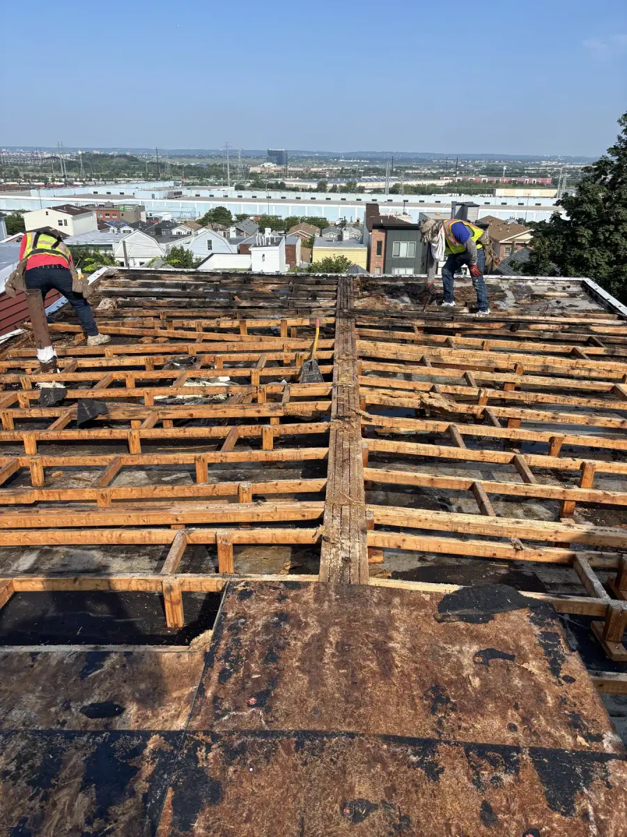 A roof stripped back to bare rafters with two crew members working across it and the city beyond